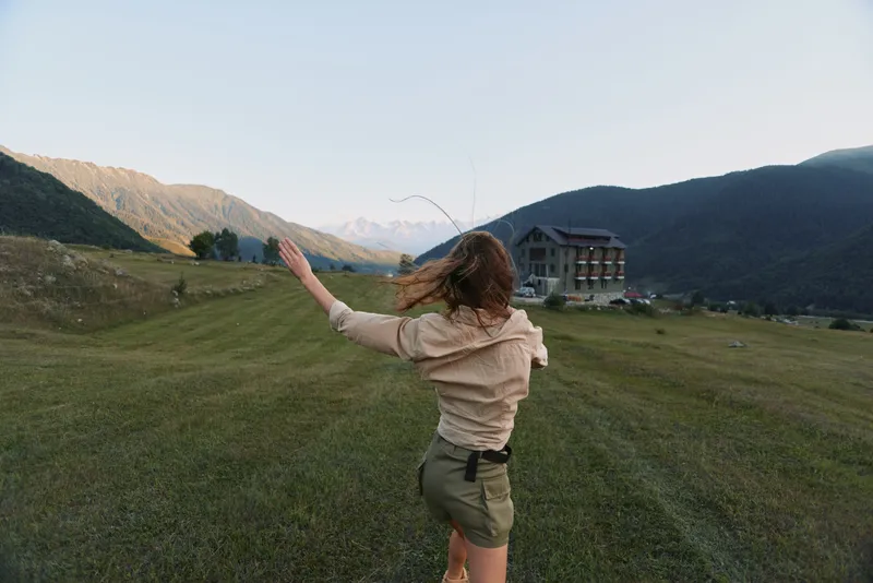 A woman in casual clothing runs through a grassy hillside, arms outstretched, embracing wide open nature. A mountainous landscape frames the bright, peaceful countryside.