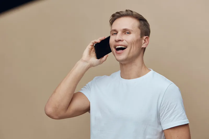 Smiling young man with short brown hair in white t-shirt talking on smartphone isolated on beige background studio portrait. People lifestyle concept
