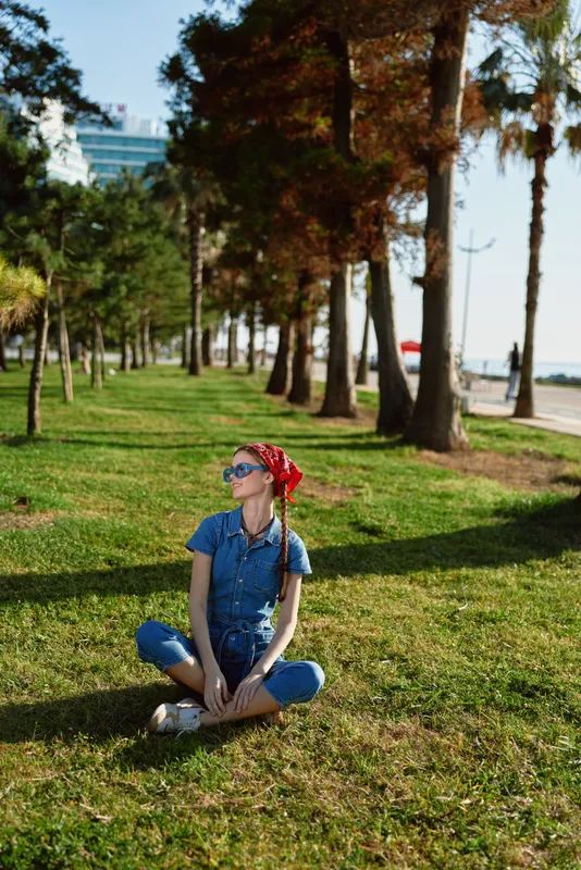 young woman sitting on grass under palm trees in sunny park wearing sunglasses and casual blue outfit enjoying warm weather and relaxing outdoors
