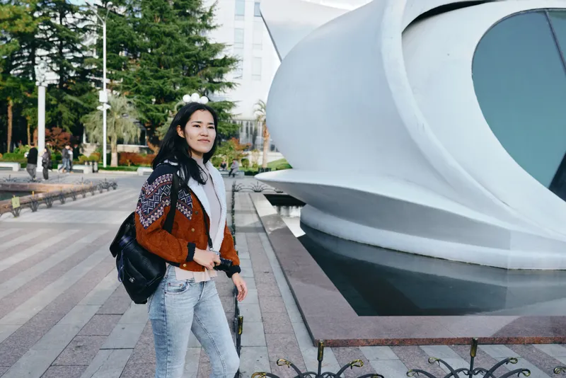 Woman with backpack walking beside futuristic architecture on urban street, exploring city scenery, casual fashion, travel vibe, candid moment near modern sculpture