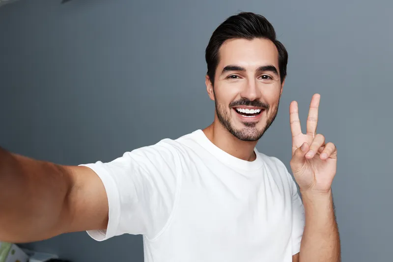 Smiling man making peace sign with hand taking selfie in bright casual t-shirt against simple gray background. Joyful expression and cheerful mood concept.