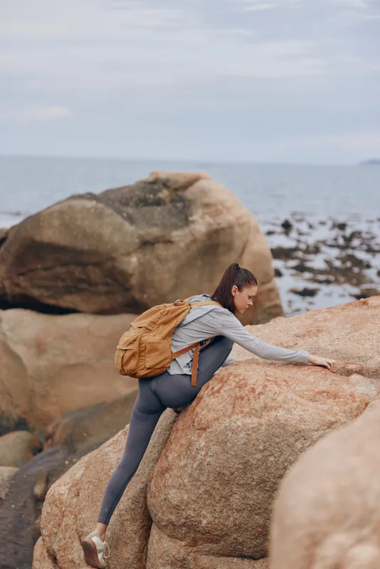 A young woman climbing rocky terrain by the sea, showcasing determination and adventure in a casual outfit, perfect for outdoor enthusiasts seeking inspiration