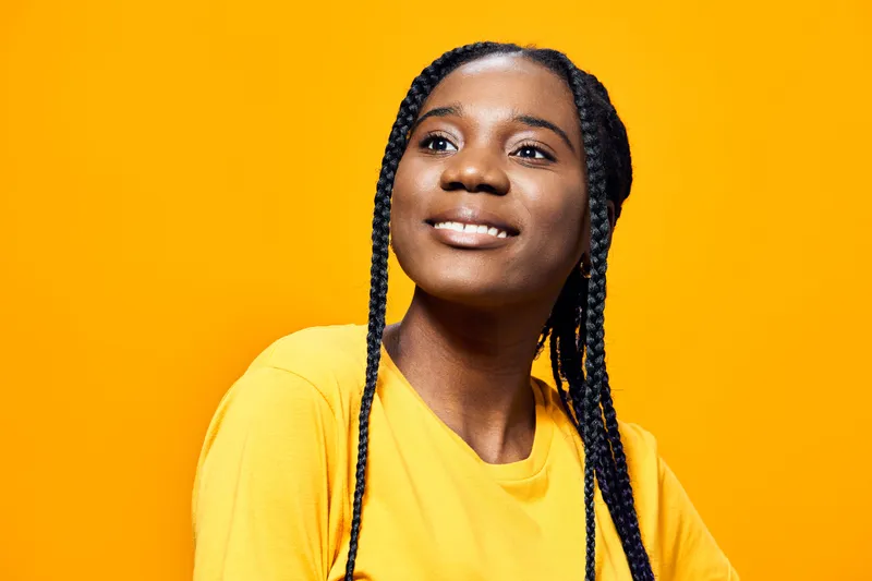 Smiling young woman with braided hair wearing a yellow t shirt against a bright orange background, conveying joy and positivity in a vibrant, cheerful atmosphere