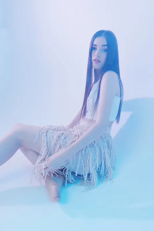 Young woman with long hair wearing a fringed skirt and white top sitting on a smooth surface. Soft blue lighting highlights fashion and beauty concepts in a studio setting.