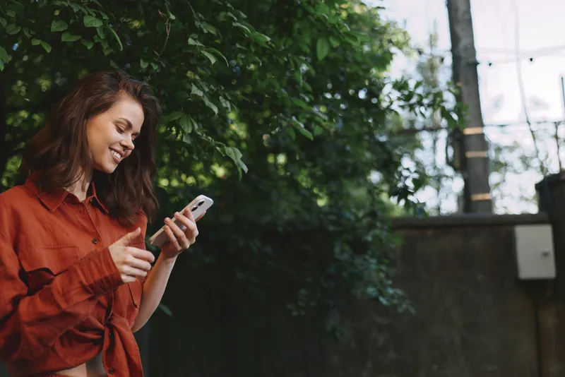 A woman in a red blouse stands outdoors, smiling while looking at her phone, capturing a casual lifestyle moment with greenery and bright daylight