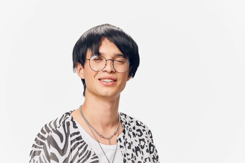 smiling man wearing round glasses and patterned shirt with layered silver necklaces, joyful expression against plain white background, young adult, casual style