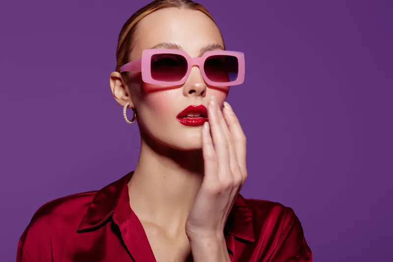 Stylish woman with pink sunglasses and red lipstick poses against purple background. Fashion portrait with trendy accessories and confident expression. Concept of glamour and modern style.
