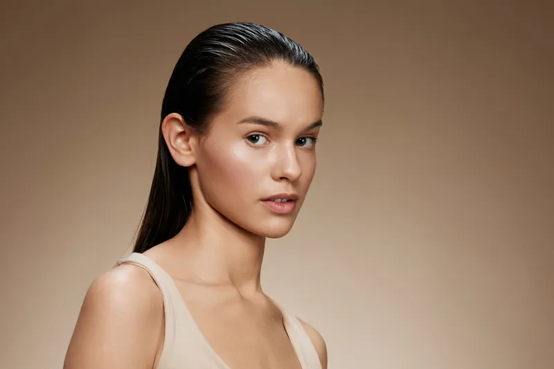 Young woman with wet hair and natural makeup against a neutral background, showcasing beauty and confidence in a soft, warm color palette