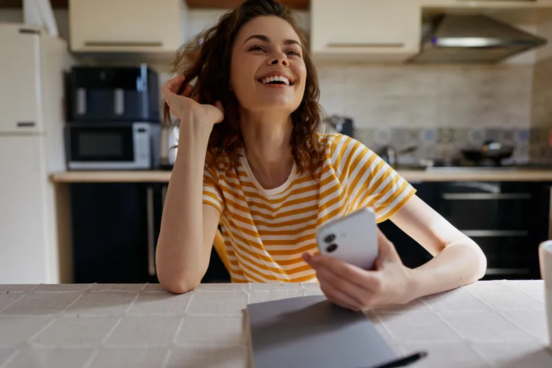 Smiling woman wearing a striped shirt sits at a home kitchen table using a smartphone, showcasing casual lifestyle, cozy indoor ambiance, and confident self expression and genuine warmth.