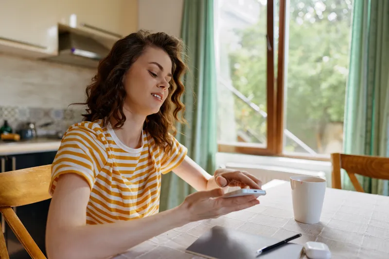 Woman in a striped shirt sits at a kitchen table at home, using a smartphone with a coffee cup nearby, conveying a casual morning lifestyle and relaxed mood.