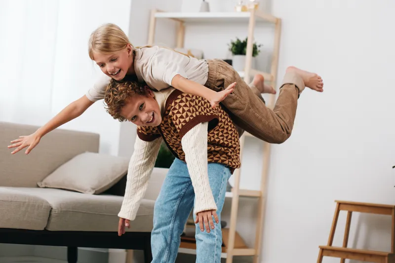 Authentic moment of children at home, a playful scene as one child carries another on a sofa, capturing genuine joy, energy, and everyday lifestyle.
