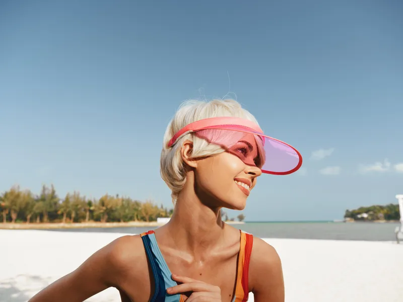 Sunlit beach portrait featuring a smiling woman in a pink visor and sporty swimsuit, with bright blue sky and calm sea backdrop, capturing carefree summer energy.