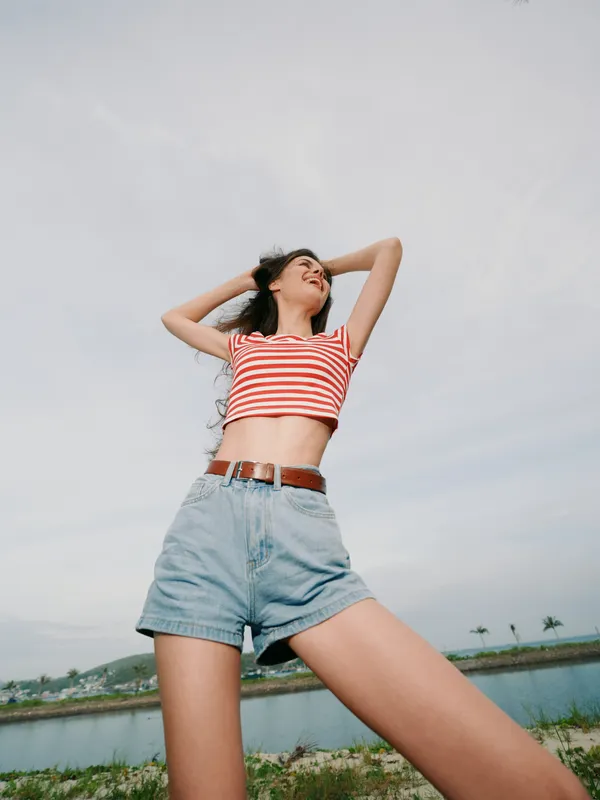 Young woman stands outdoors by a lake, wearing a red and white striped crop top and denim shorts, arms raised, head back, capturing carefree summer energy.