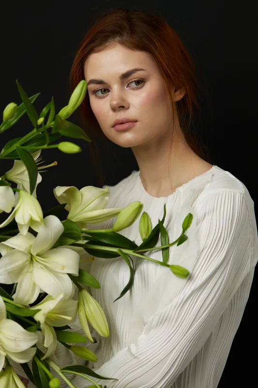 Young woman with red hair holding white lilies, wearing a light textured blouse, exuding a calm and serene emotion against a black background, perfect for floral and lifestyle themes