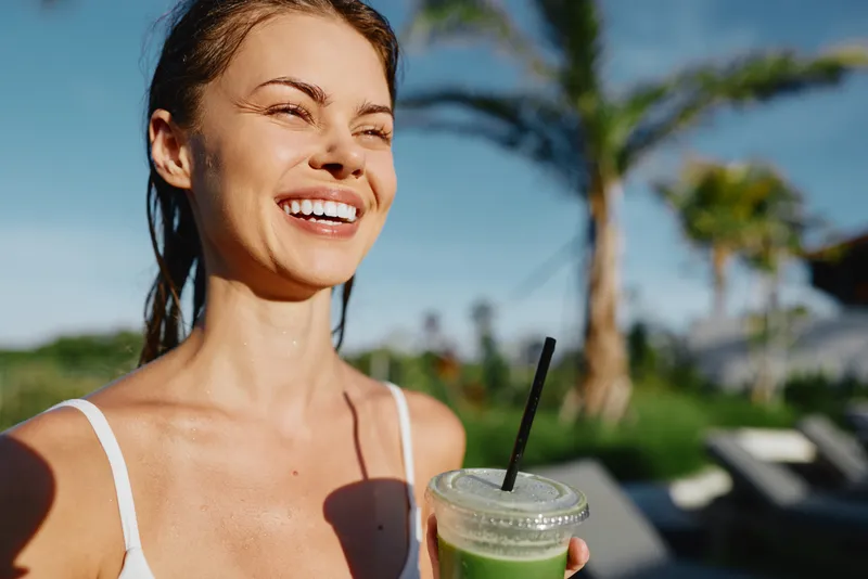 Happy woman outdoors smiling with eyes closed holding green smoothie cup, sunny day, casual white top, relaxed lifestyle, palm trees and blue sky background