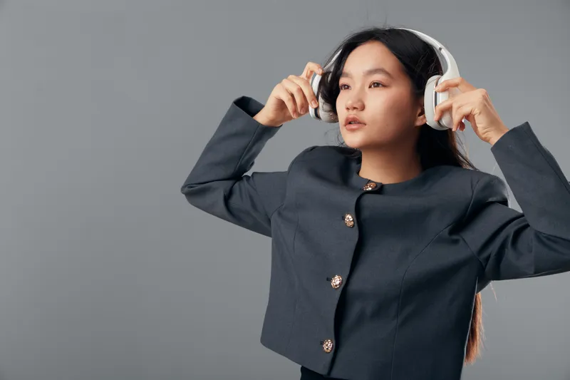 Young woman wearing dark blouse adjusting wireless headphones on gray background for music listening, technology, entertainment, and modern style with focused expression in studio.