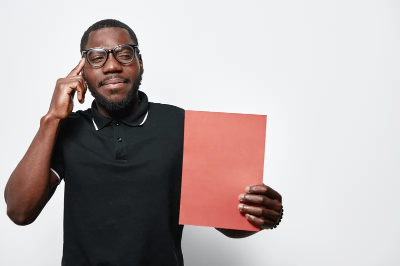 African American man with dark skin and glasses holding red paper and pointing to head for idea or thinking concept on white background.
