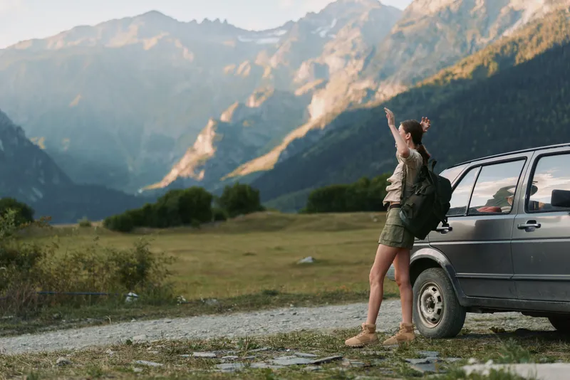 Woman with backpack standing near car on mountain road pointing at distant peaks. Outdoor lifestyle, adventure, travel, and nature exploration concept in warm daylight.