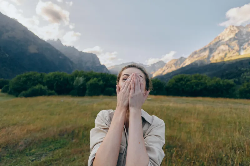 A young woman with brunette hair covers her face with her hands in a meadow as distant mountains rise behind her, expressing surprise and wonder.