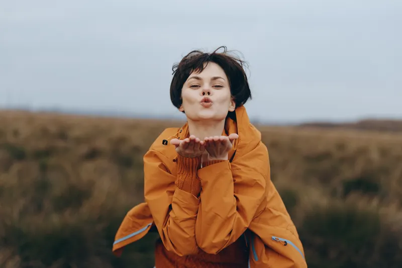 Woman lifestyle outdoors field autumn wind portrait orange jacket casual, young woman in orange coat in open countryside, wind moves hair, soft daylight, happy mood, calm nature background