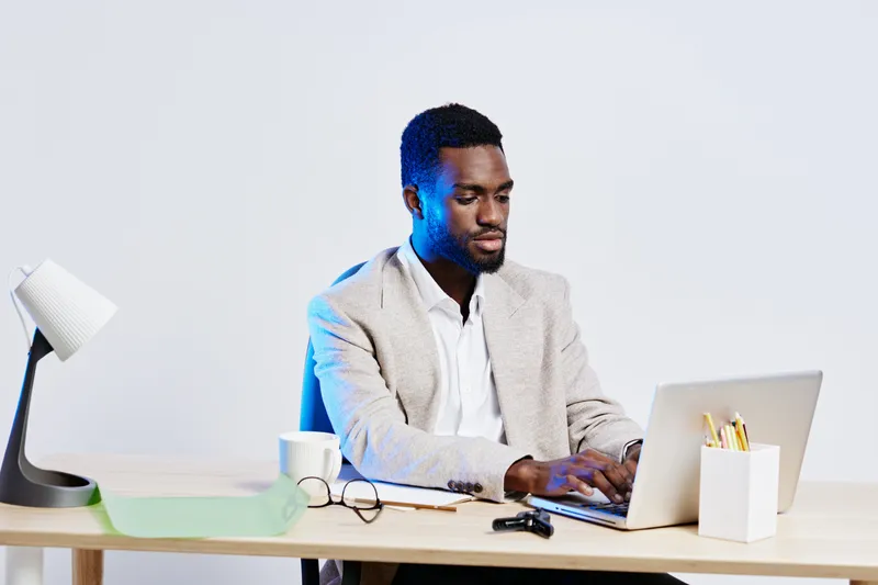 Professional young man working at modern desk using laptop, wearing business casual attire, focused on screen in bright office environment, supplies and accessories nearby. People lifestyle concept