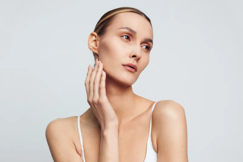 Young woman with clear skin gently touches her face in a soft studio setting, conveying calm and self care.