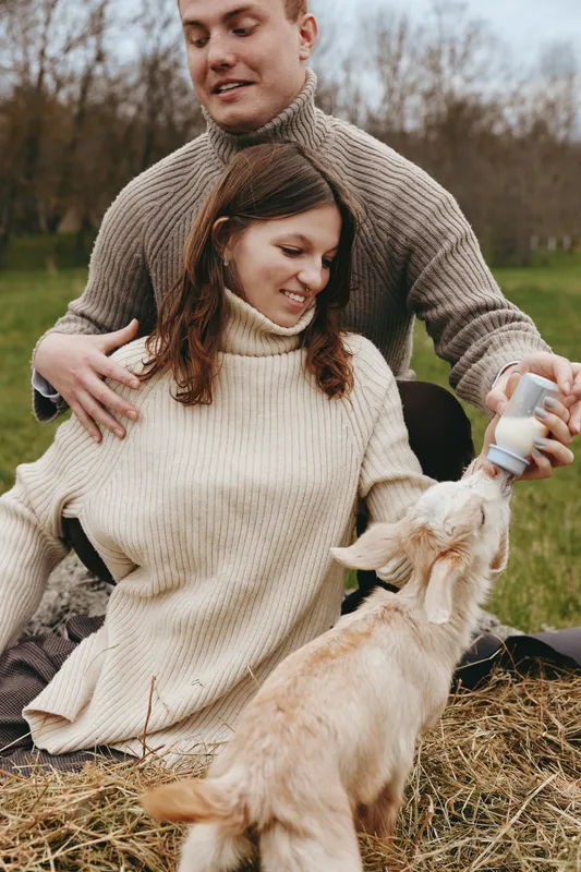 Couple outdoors share a warm moment with a goat kid, gentle feeding, cozy sweaters, natural colors, sunlit field, calm interaction, affectionate scene, relaxed lifestyle photography