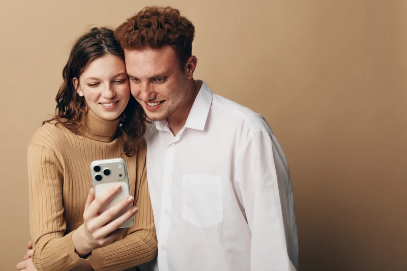 Smiling young couple takes selfie with smartphone, standing close together on beige background. Concept of friendship, technology, happiness, and casual lifestyle in modern relationships.