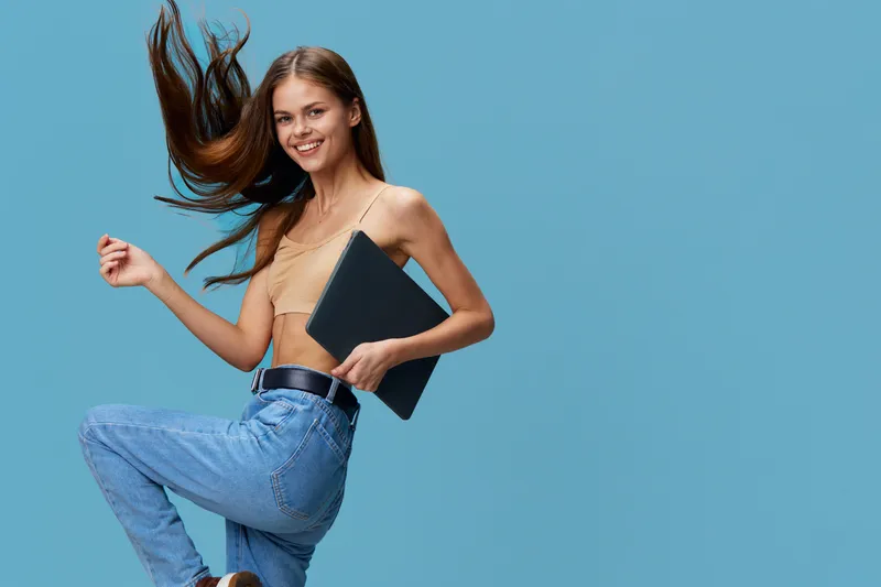 smiling woman holding laptop jumping with joy wearing casual jeans and crop top on blue background, reflecting happiness and youthful energy