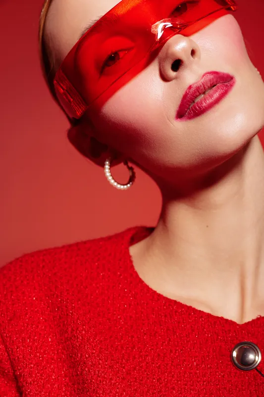 Close-up portrait of a woman wearing red sunglasses and a red textured top with a button, posing against a red background. Concept of fashion, style, and modern accessories.