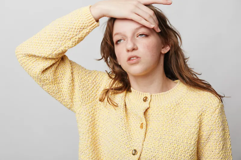 Tired young woman in yellow knitted sweater touches forehead with hand, showing fatigue and discomfort. Isolated on light background. Concept of exhaustion, headache, and stress relief.