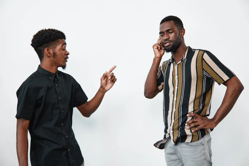 Two African American men with dark skin having a serious conversation, one pointing finger and the other listening on phone, casual clothing on white background.