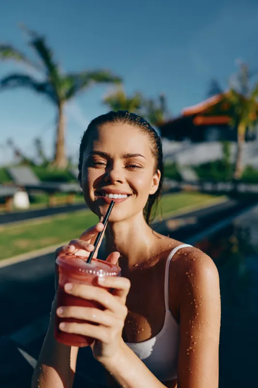 Smiling woman in a white tank top sipping a pink smoothie beside a sunny pool in a tropical outdoor setting, showcasing refreshment, wellness and carefree summer vibes.