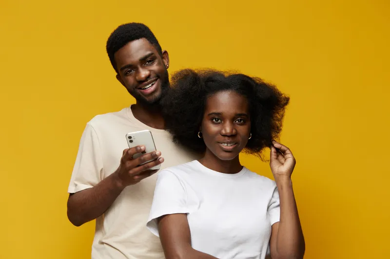 Happy young Black couple smiling together against a vibrant yellow background, showcasing casual fashion and a joyful atmosphere while using a smartphone