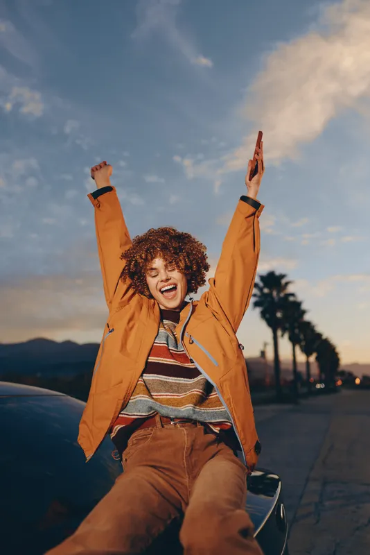 Woman lifestyle outdoors at sunset, curly haired young woman smiling on scooter, raising both arms, wearing brown jacket and striped top, street background evening city lights.