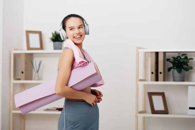 Woman in athletic wear smiles while wearing headphones and carrying a pink yoga mat in a bright home or office space, ready for a workout and fitness routine.