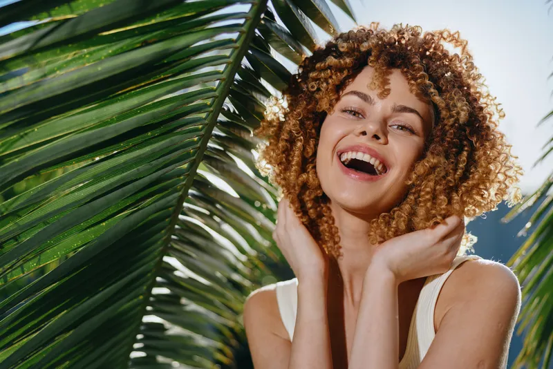 smiling woman with curly hair poses happily near tropical palm leaves in bright natural sunlight, wearing a white sleeveless top outdoors in summer.