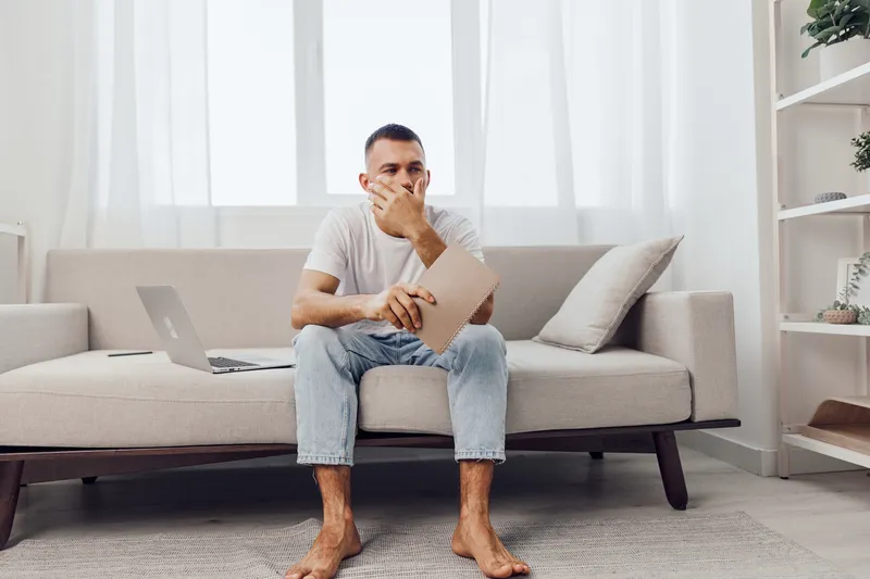 Thoughtful man sitting on a cozy sofa, holding a notebook and looking contemplative, surrounded by a bright, modern living space that reflects relaxation and focus.