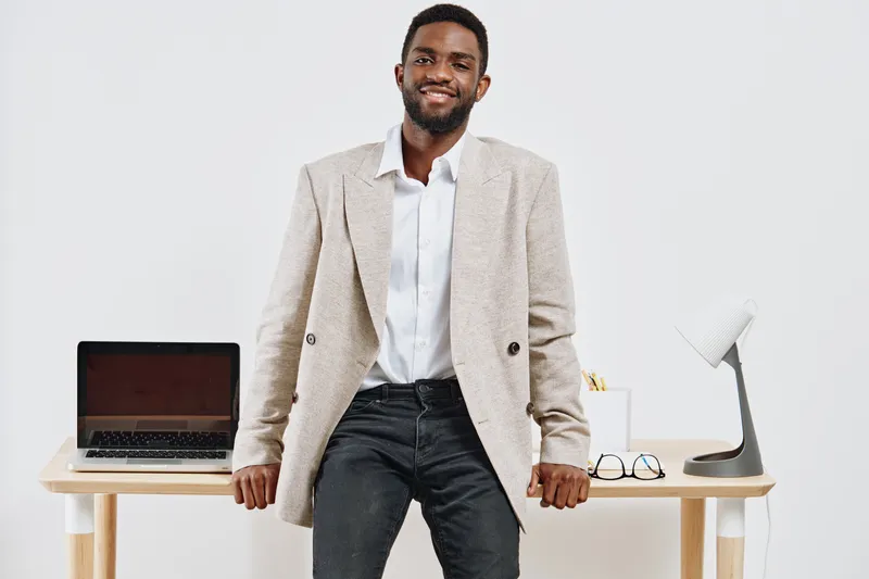 Confident smiling man standing in modern office corridor, casual beige blazer, white shirt, jeans, relaxed pose, professional yet approachable appearance, bright minimalistic background, lifestyle con