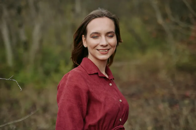A candid outdoor portrait of a smiling young woman in a burgundy dress, conveying natural expression and subtle authenticity and credibility in a relaxed woodland setting, warm tones and soft light.