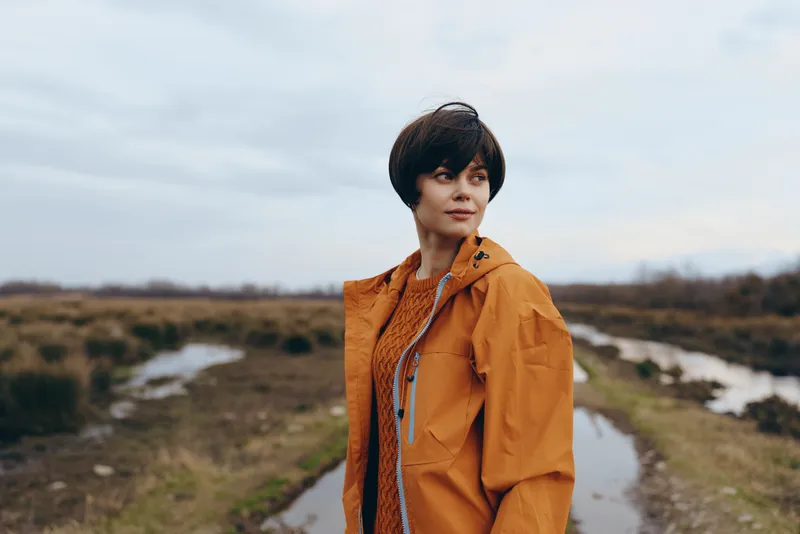 Woman lifestyle outdoors portrait in orange jacket, standing on rural path, looking at camera, soft overcast light, gentle breeze, natural colors, blurred hills and shrubs behind