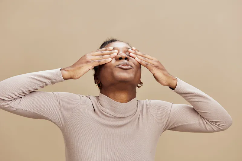 Emotional African woman gently touching her face against a warm beige background, expressing relaxation and calmness with a serene smile