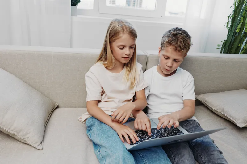 Two children sitting on a sofa using a laptop together in a bright room, focused on the screen. Concept of learning, technology, childhood, and collaboration.