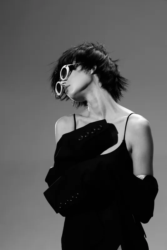 Black and white portrait of a stylish woman in sunglasses wearing a black outfit with a modern edgy look.