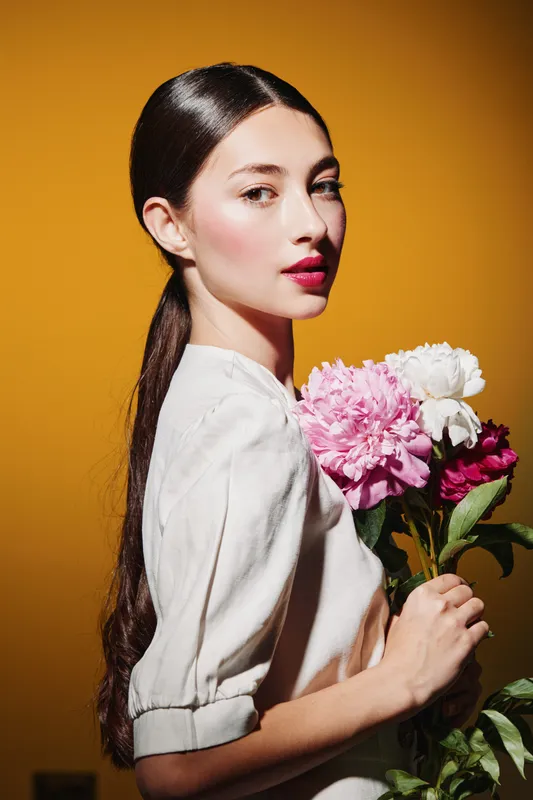 Elegant woman with sleek dark hair and radiant skin holding colorful vibrant flowers on a warm yellow background. Concept of beauty, fashion, and creative floral portrait.