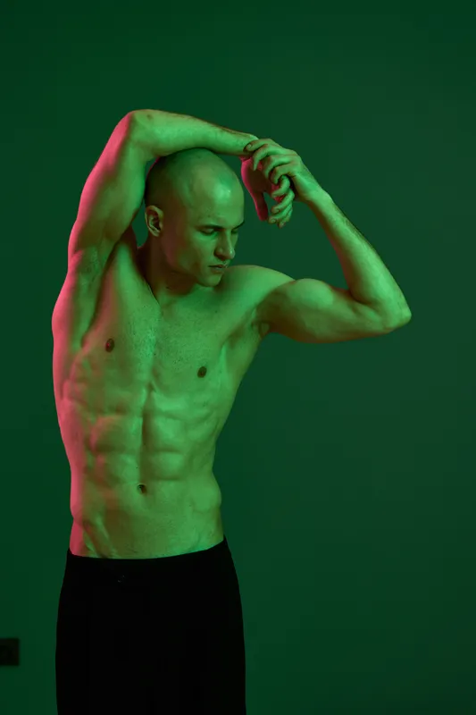 Athletic man posing with dramatic lighting in a green and pink hue, showcasing strength and elegance. This artistic photo captures unique body lines and emotion.