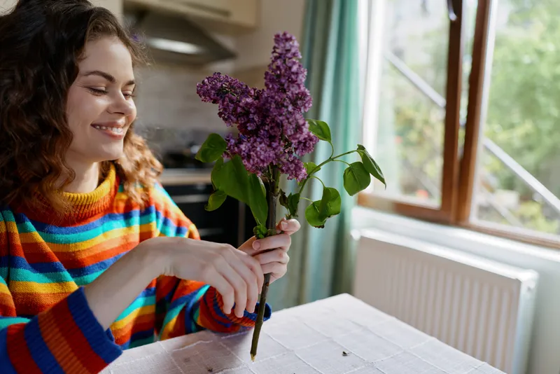 Cheerful woman indoors, arranging a bouquet of lilac flowers, bright striped sweater, warm natural light, cozy kitchen setting, relaxed mood, everyday home life