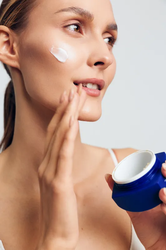 Young woman applying moisturizing cream on her cheek with a blue jar in hand. Close-up portrait with natural skin, skincare routine, and beauty concept on white background.