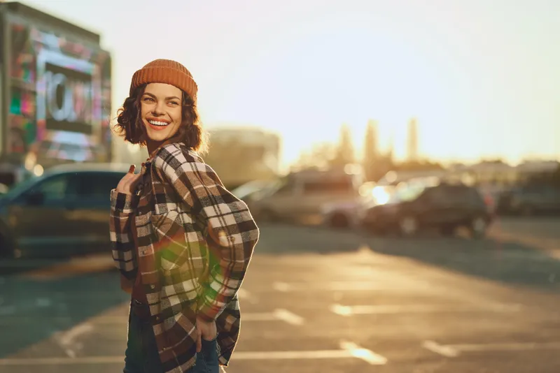 woman smile urban sunset beanie plaid candid golden authenticity mindful living emotional storytelling. Smiling young woman in a city parking lot at golden hour glow, warm sunlight and relaxed