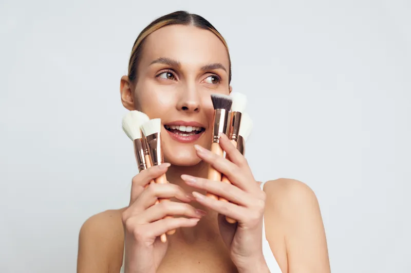 Woman beauty portrait closeup on white background, holding cosmetics containers near face, smiling and carefully applying makeup, elegant spa skincare routine for youthful fresh skin.